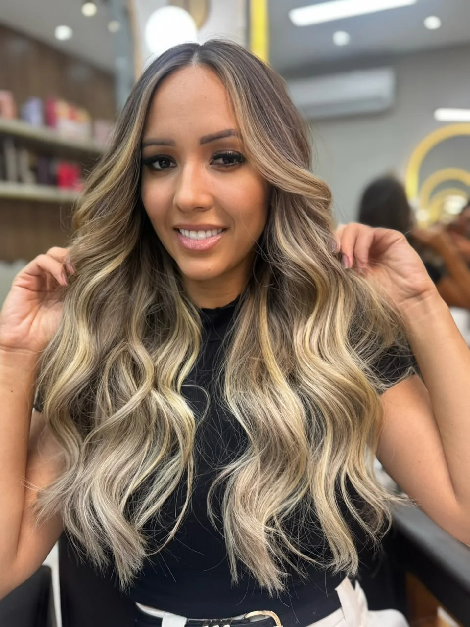 Cabelos longos com mechas loiras e ondas, com a cliente segurando as laterais do cabelo